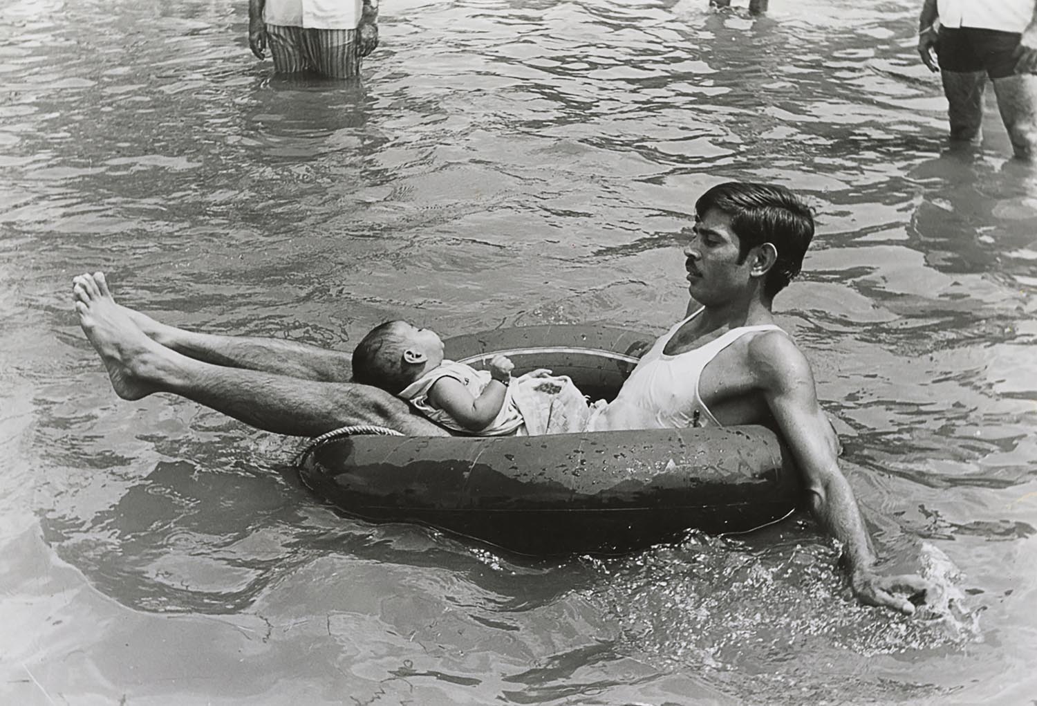 A man on a rubber float with a baby on his lap, carrying themselves to safety during a flood.